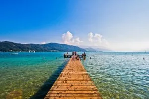 Ein Holzsteg ragt in einen klaren, türkisfarbenen See hinein, auf dem Menschen stehen und sitzen; Schwimmer sind im Wasser und im Hintergrund sind Berge unter einem strahlend blauen Himmel mit flauschigen Wolken zu sehen.
