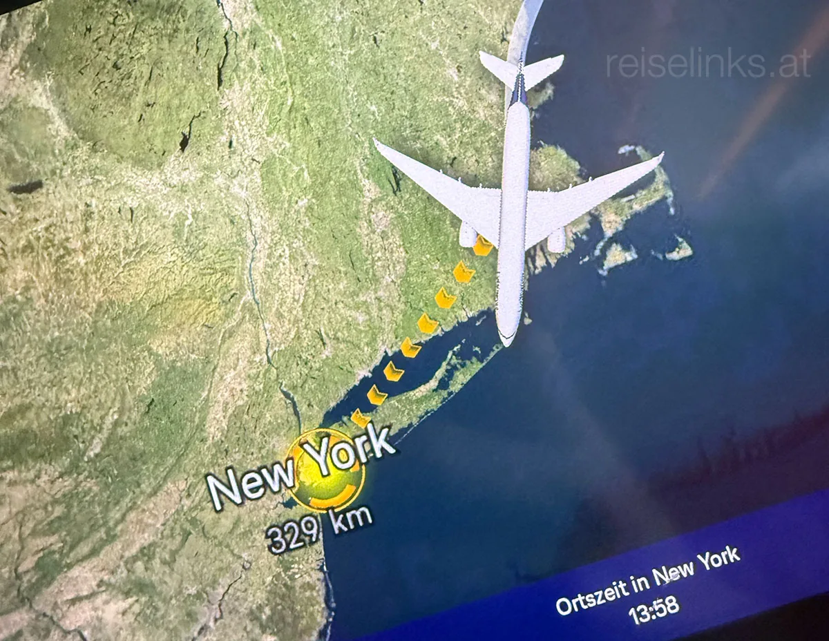 Eine digitale Flugkarte zeigt ein Flugzeug im Anflug auf New York (329 km Entfernung). Eine gestrichelte gelbe Linie markiert den Weg. Unten wird die Ortszeit New Yorks (13:58 Uhr) angezeigt.