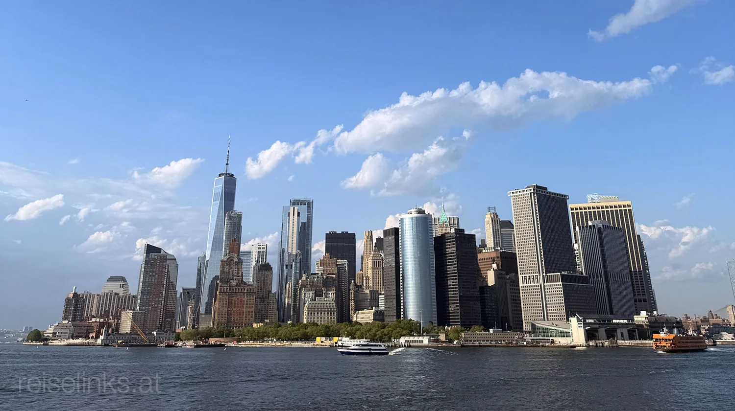 Die Skyline von Manhattan an einem sonnigen Tag vom Wasser aus betrachtet, mit hohen Wolkenkratzern, darunter das One World Trade Center, unter einem blauen Himmel mit vereinzelten Wolken. Im Vordergrund sind Boote auf dem Fluss zu sehen.
