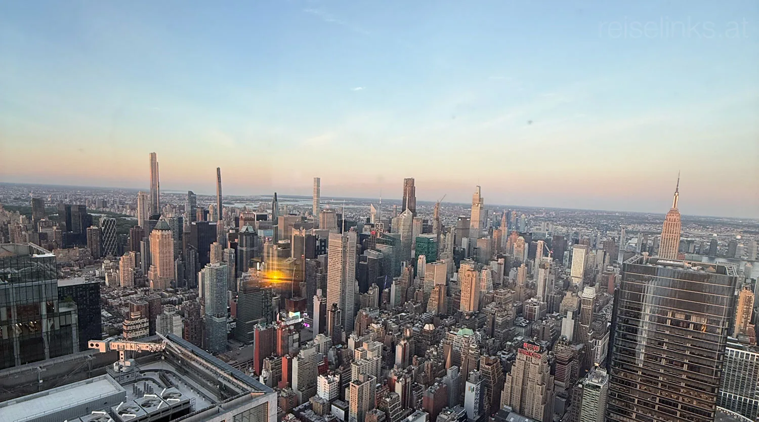 Luftaufnahme der Skyline von New York City bei Sonnenuntergang mit zahlreichen hohen Wolkenkratzern, darunter das Empire State Building auf der rechten Seite, unter einem klaren Himmel, in dem sich das sanfte Sonnenlicht in den Gebäuden spiegelt.