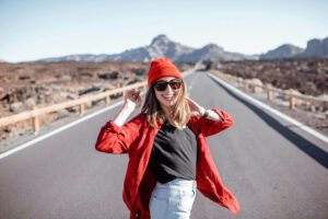 Eine junge Frau mit rotem Hut, roter Jacke, schwarzem Hemd und Sonnenbrille steht lächelnd auf einer leeren Straße mit Bergen im Hintergrund unter einem klaren Himmel.